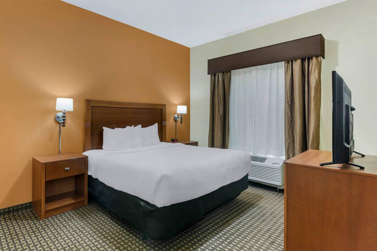 A hotel room with a large bed, two bedside tables with lamps, a flat-screen TV on a wooden stand, and an air conditioning unit.