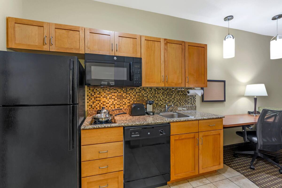 A compact kitchen with wooden cabinets, a black refrigerator, a microwave, a dishwasher, and a tiled backsplash.
