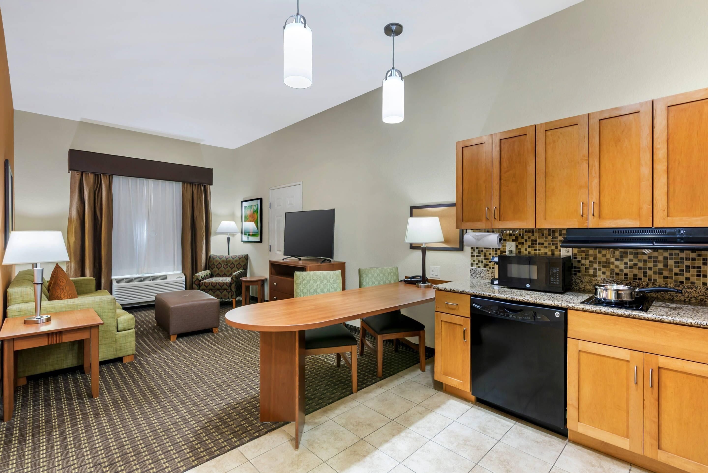 A hotel room with a kitchenette, a dining area, a living room with a sofa and a TV, and a window with curtains.