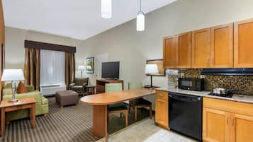A hotel room with a kitchenette, a dining area, a living room with a sofa and a TV, and a window with curtains.