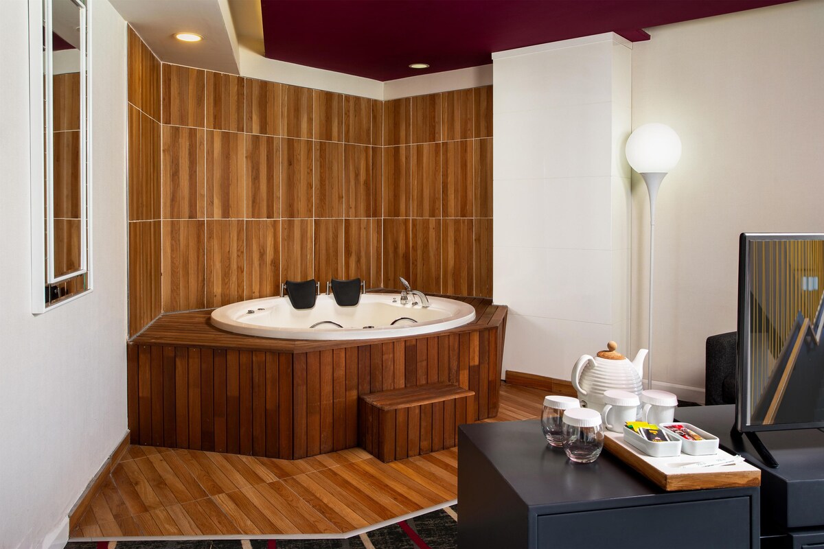 Un baño moderno con pared de panel de madera, una bañera independiente, un tocador con espejo y una mesita de noche con una lámpara y una tetera.