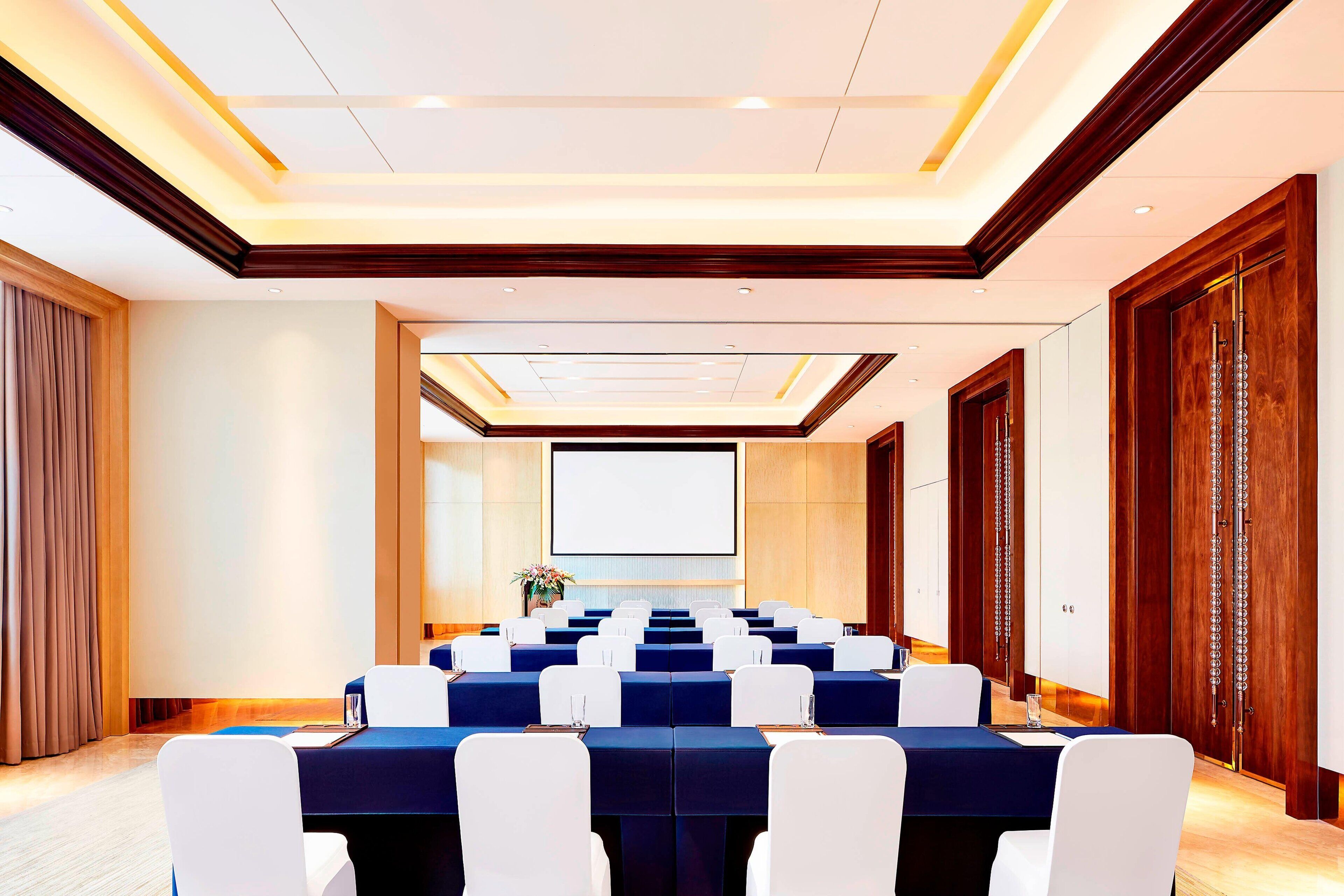 A conference room with a long table, white chairs, and a projector screen.