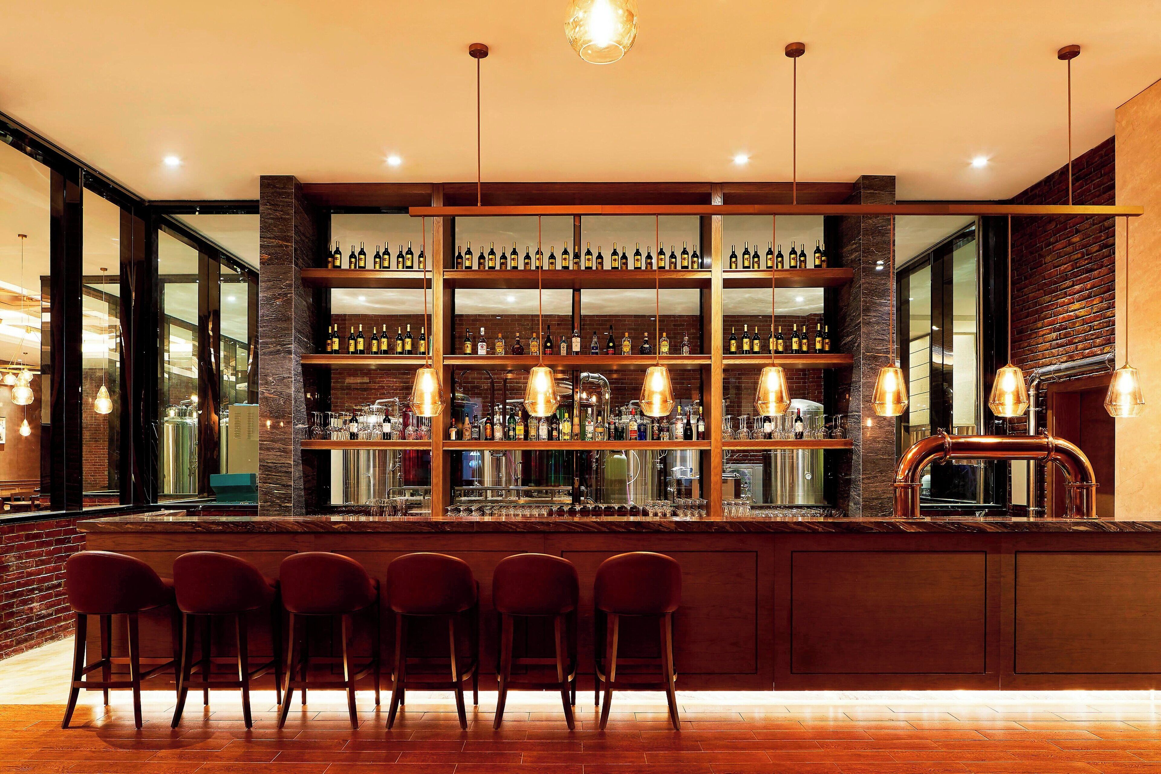 Zona de bar con varios estantes llenos de botellas, una tubería de cobre y tres taburetes de bar.