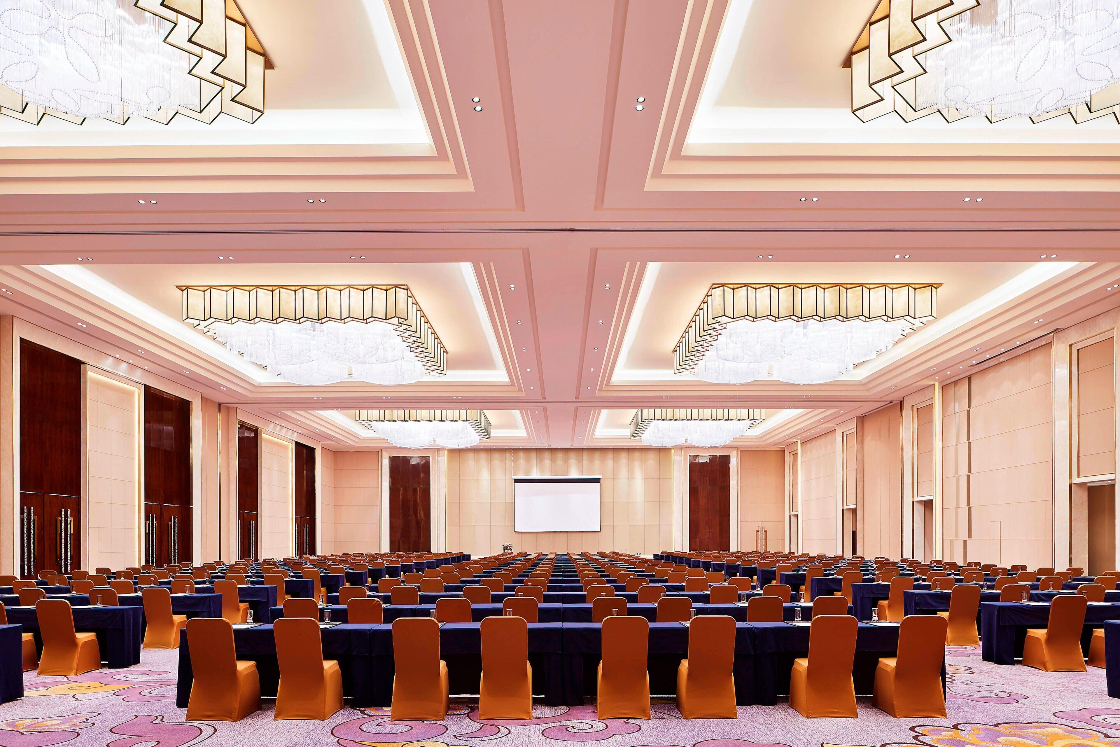 A conference room with rows of tables and chairs, a large screen at the front, and a chandelier.