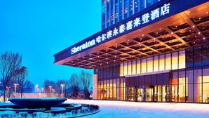 A hotel entrance with a large sign, a covered walkway, and a snow-covered ground. - Sheraton Harbin Xiangfang Hotel (Harbin)