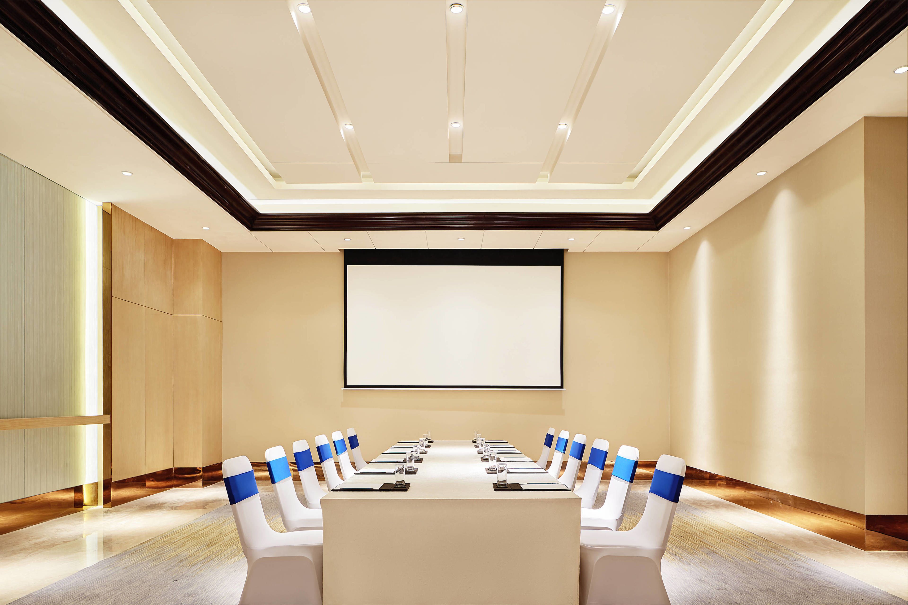 A conference room with a long table, chairs, a projector screen, and a wooden accent wall.