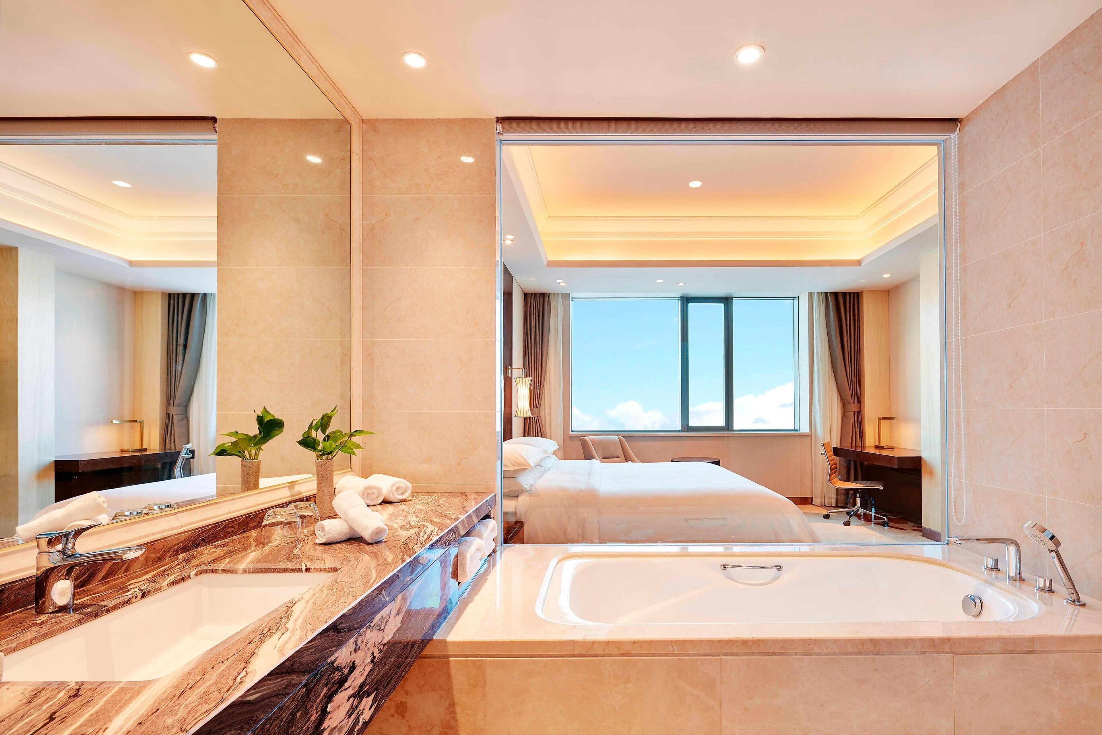 Un baño de hotel moderno con un espejo grande, una bañera, lavamanos doble y un ventanal con vistas al cielo.