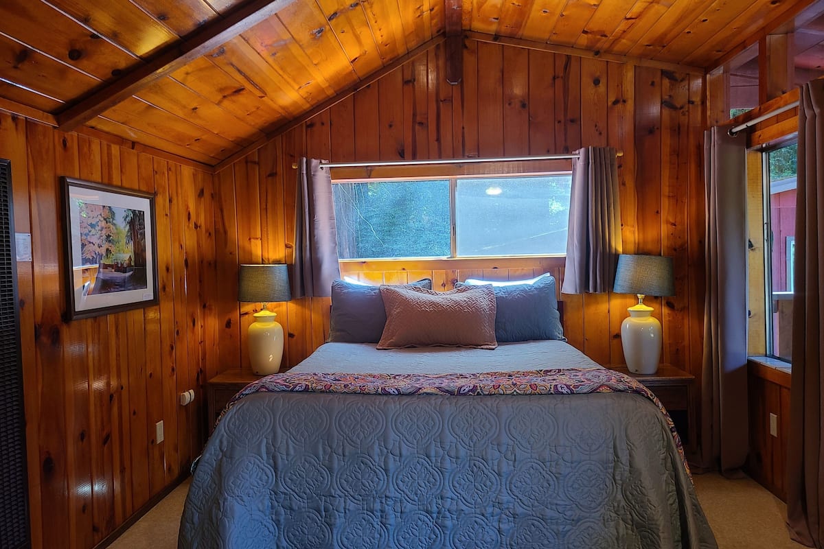 A wooden cabin bedroom with a bed, two bedside lamps, a window with curtains, and a framed picture on the wall.