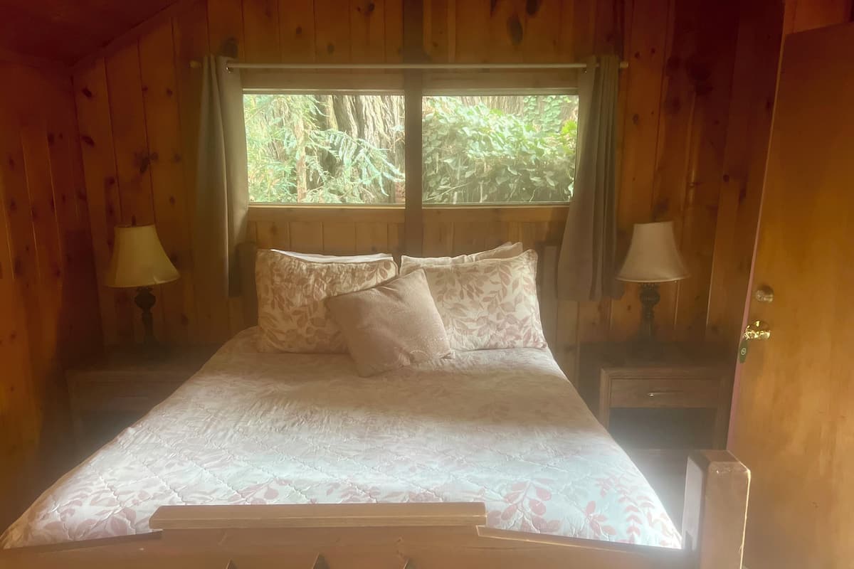 A wooden room with a bed, two bedside lamps, and a window with a view of trees.