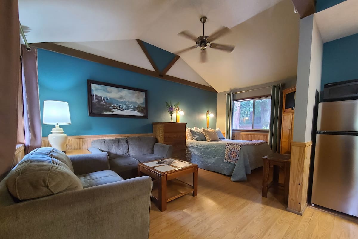 A living room with a sofa, a coffee table, a bed, a ceiling fan, and a refrigerator.