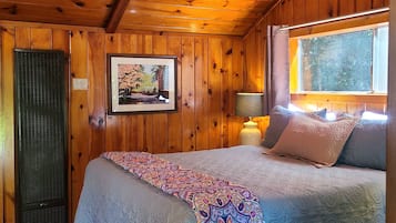 A cozy room with wooden walls and ceiling, a bed with patterned bedding, a window with curtains, and a framed picture on the wall.