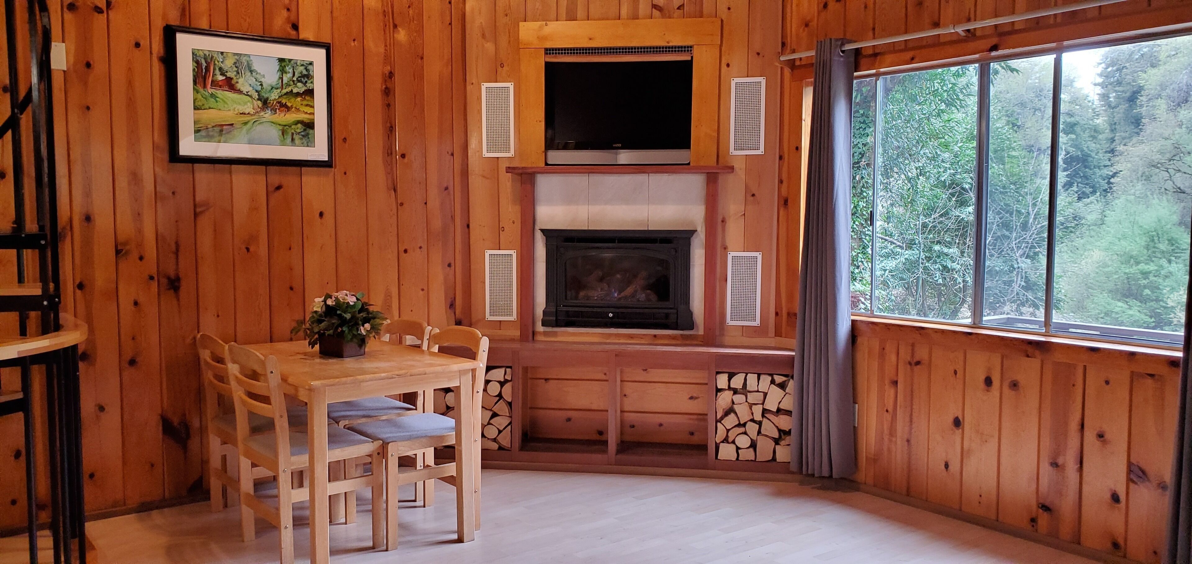 A wooden cabin interior with a fireplace, dining table, chairs, and a large window.