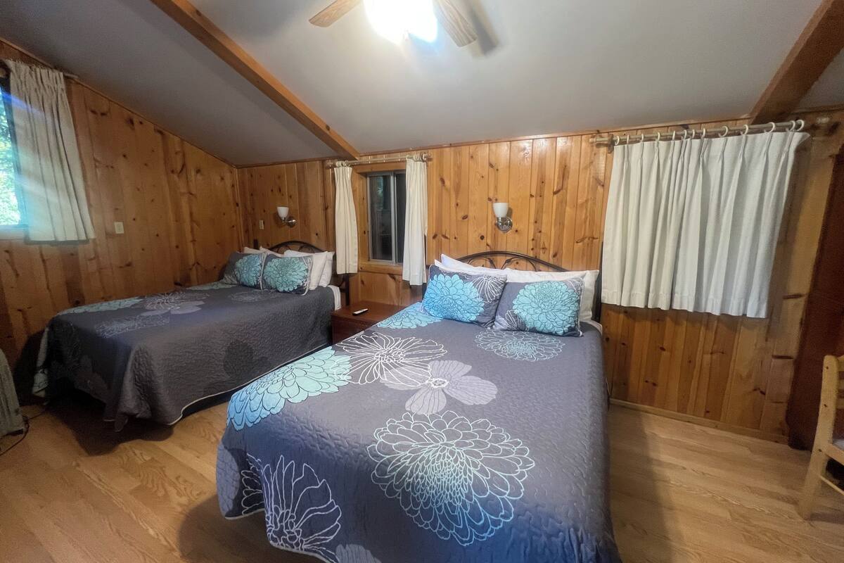 A room with two beds, wooden walls, a ceiling fan, and a window with curtains.