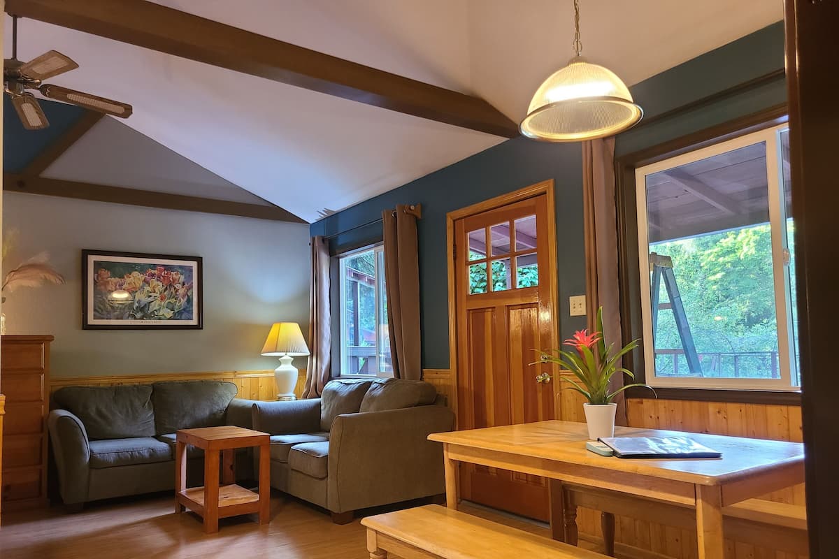 A cozy living room with a wooden bench, a small table, a sofa, and a lamp. There is a window with curtains and a painting on the wall.