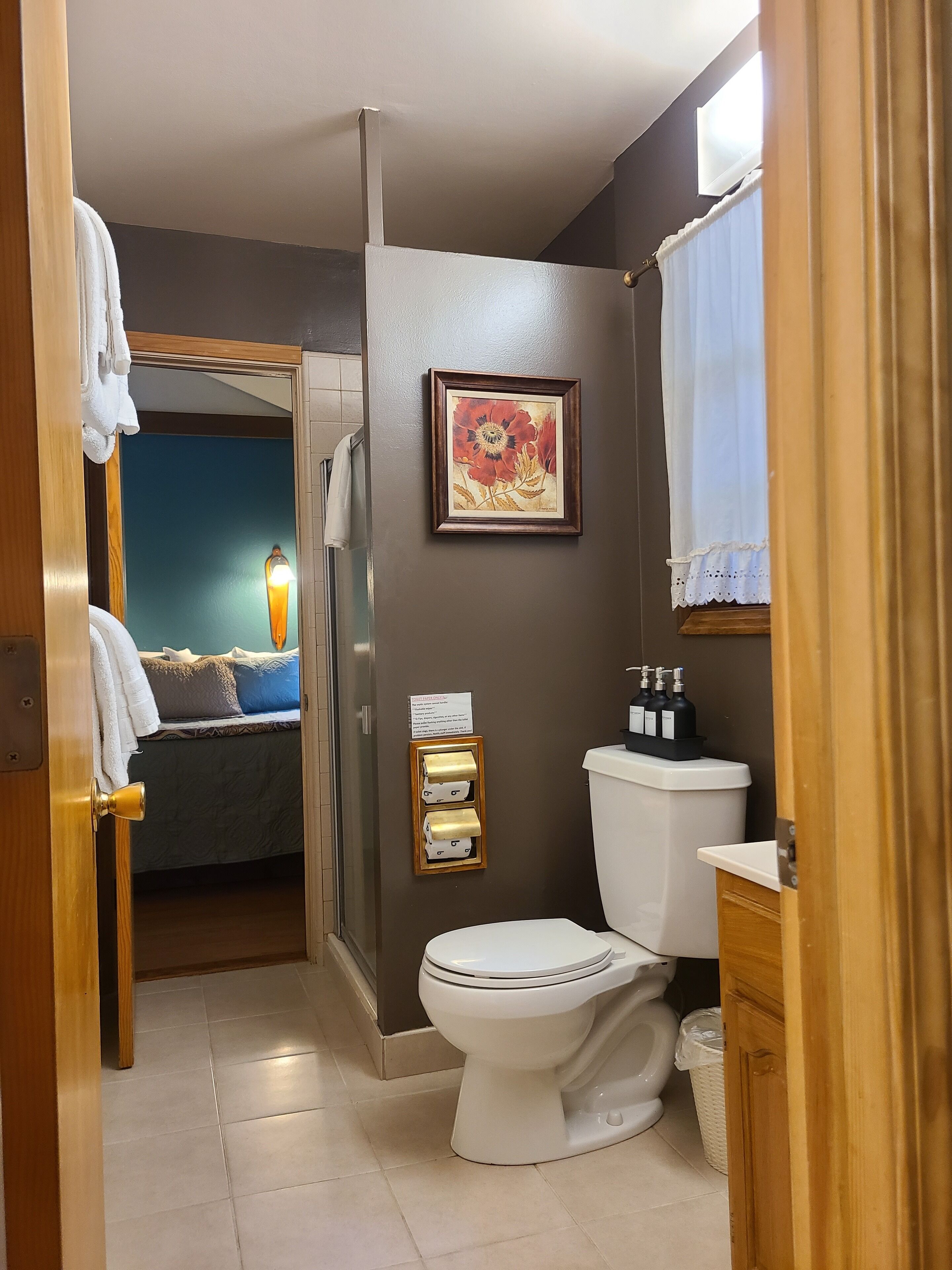 Ein Badezimmer mit Toilette, Waschbecken und Dusche. Ein gerahmtes Bild hängt an der Wand und ein Handtuch an der Tür.