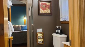 Ein Badezimmer mit Toilette, Waschbecken und Dusche. Ein gerahmtes Bild hängt an der Wand und ein Handtuch an der Tür.