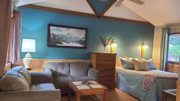 A cozy living room with a blue accent wall, a ceiling fan, a painting, a wooden cabinet, and a bed with a patterned bedspread.