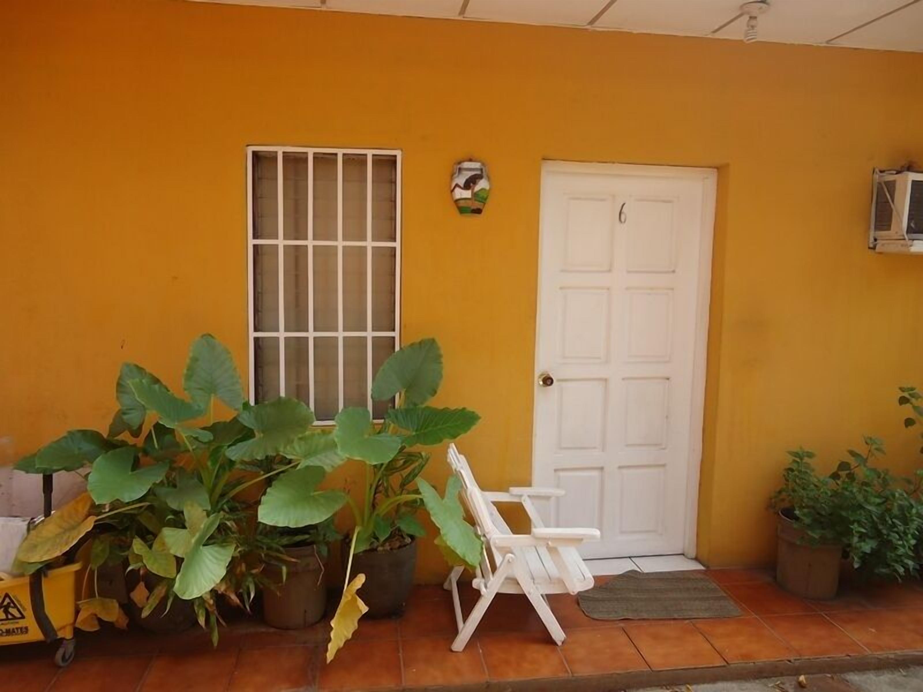 A yellow building with a white door, a window with bars, and several potted plants.