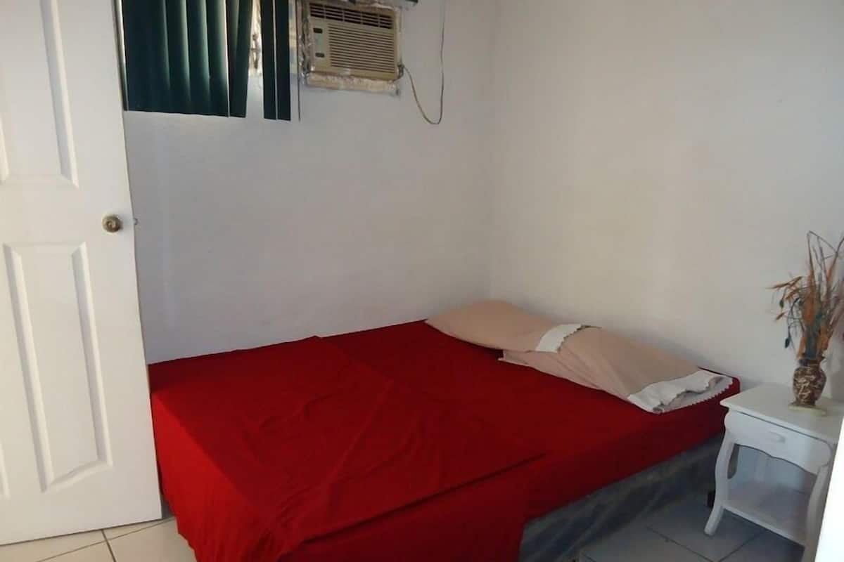 Una stanza piccola e modesta con un letto, un comodino bianco, una coperta rossa e una finestra con persiane.