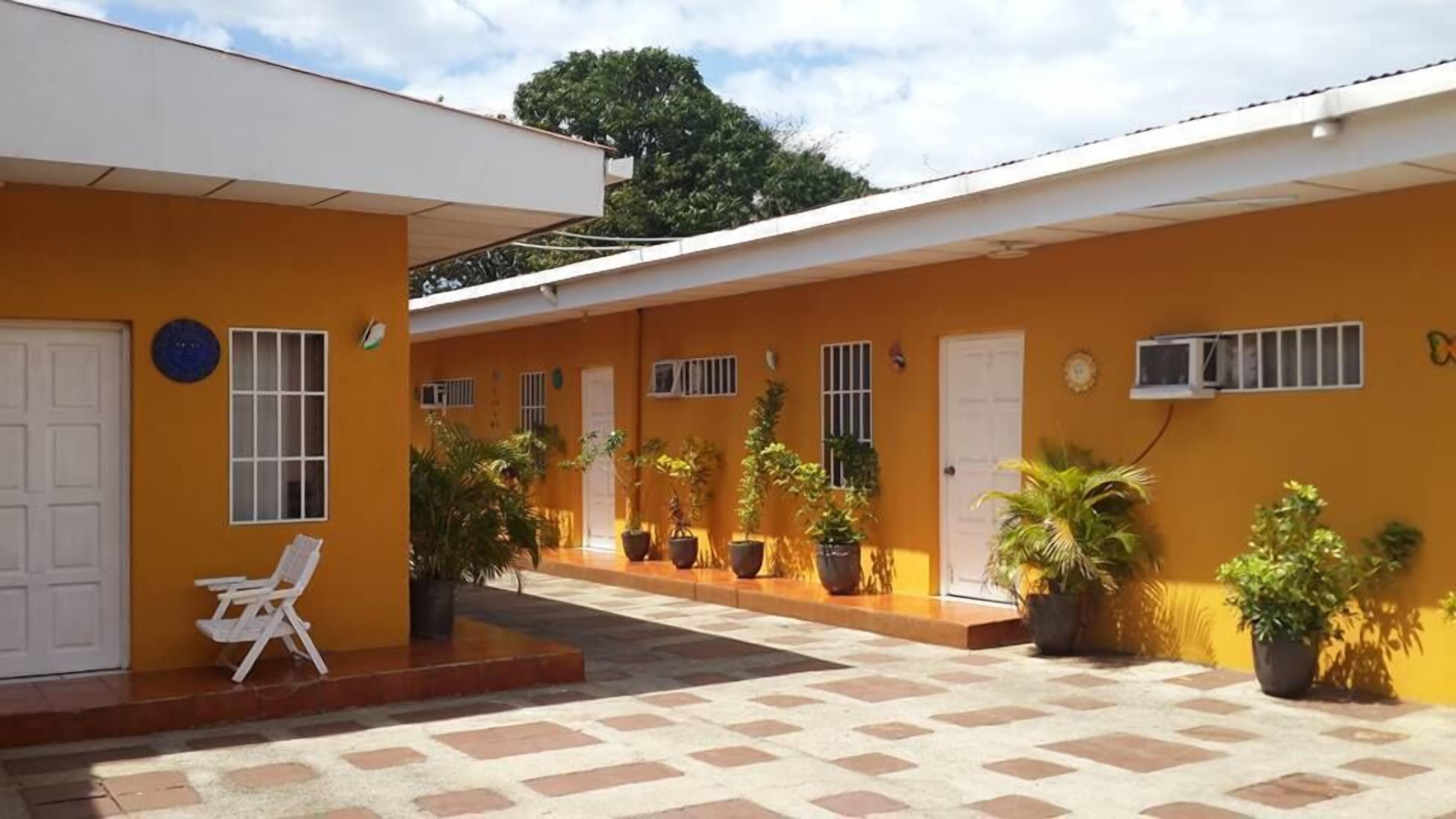 Un edificio con fachada amarilla, puertas y ventanas blancas. Hay plantas en macetas y una silla blanca afuera.
