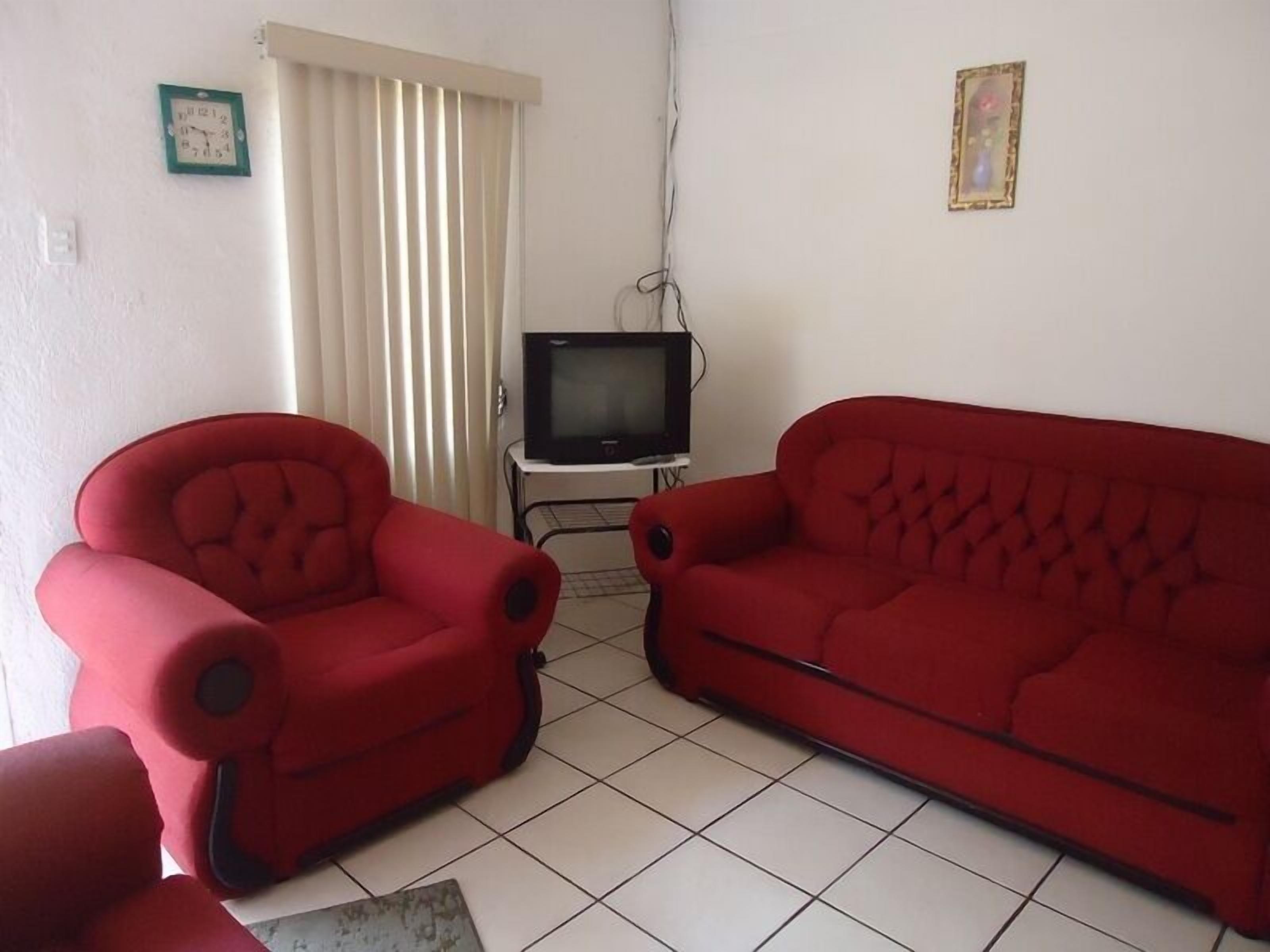 A living room with a red armchair, a red sofa, a television on a stand, and a window with vertical blinds.