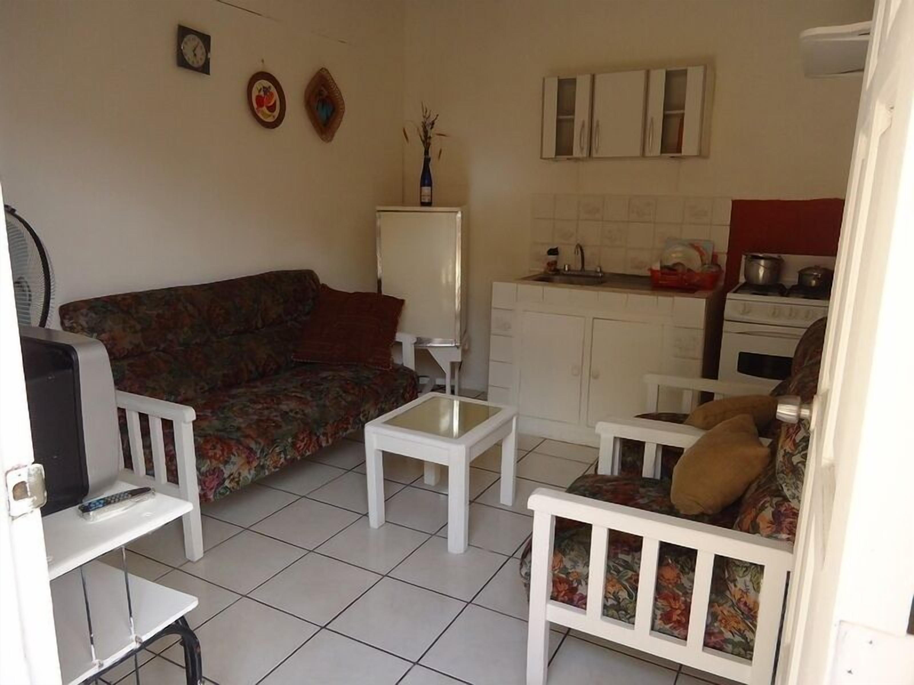A living room with a sofa, coffee table, and armchairs, a small table with a lamp, and a kitchen area with cabinets and a stove.