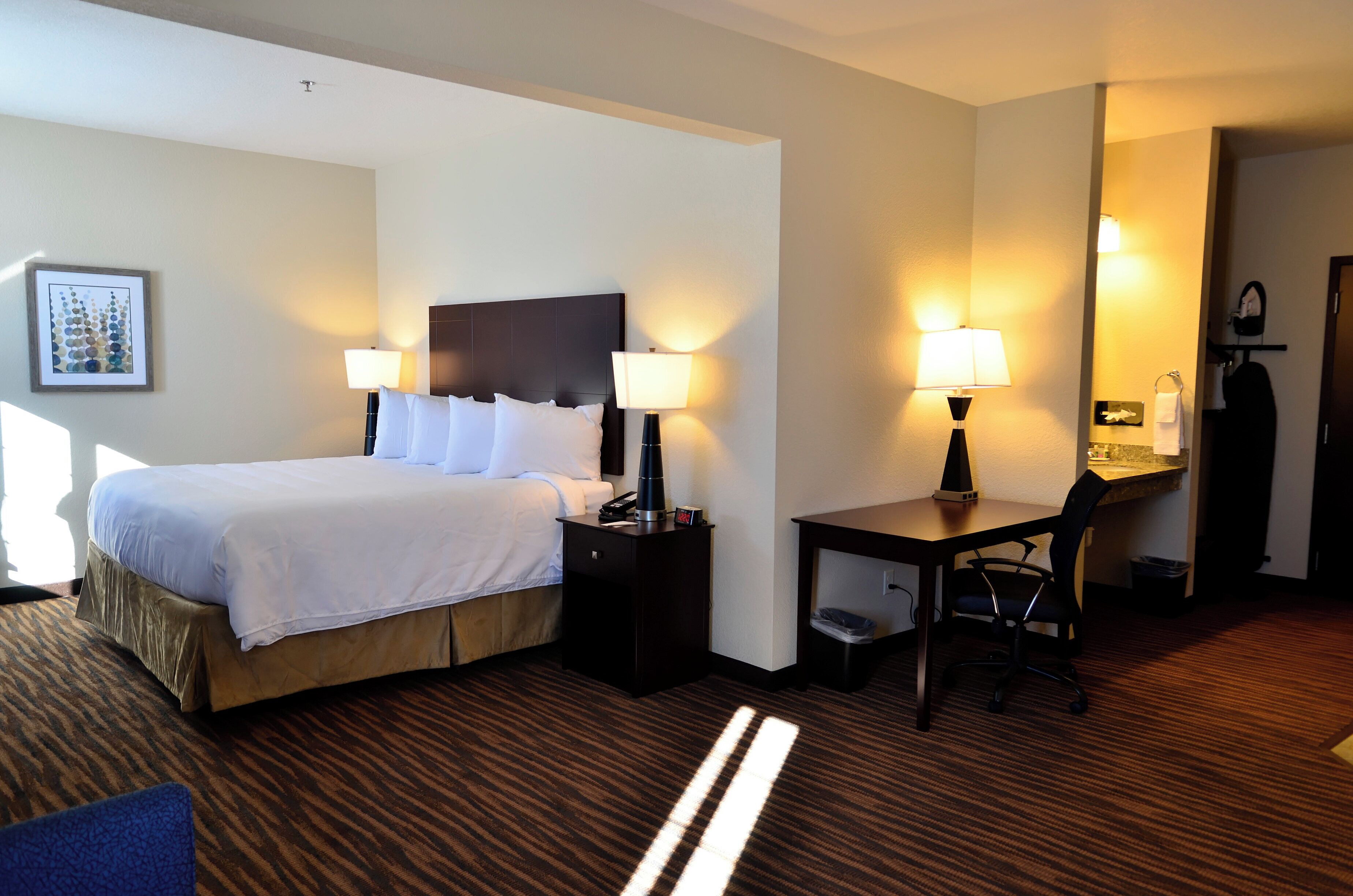 A hotel room with a bed, bedside lamps, a desk with a chair, and a small table with a lamp.