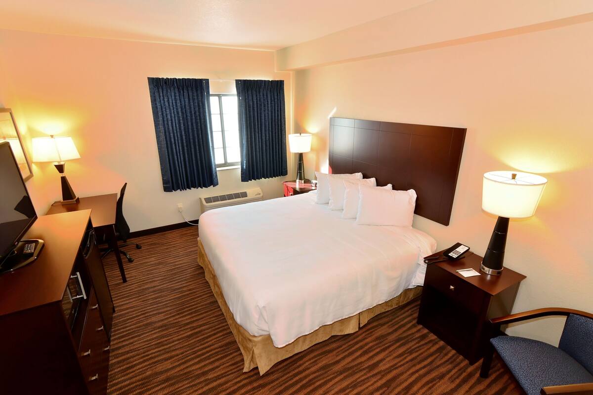 A hotel room with a large bed, two bedside tables with lamps, a desk with a chair, and a window with curtains.