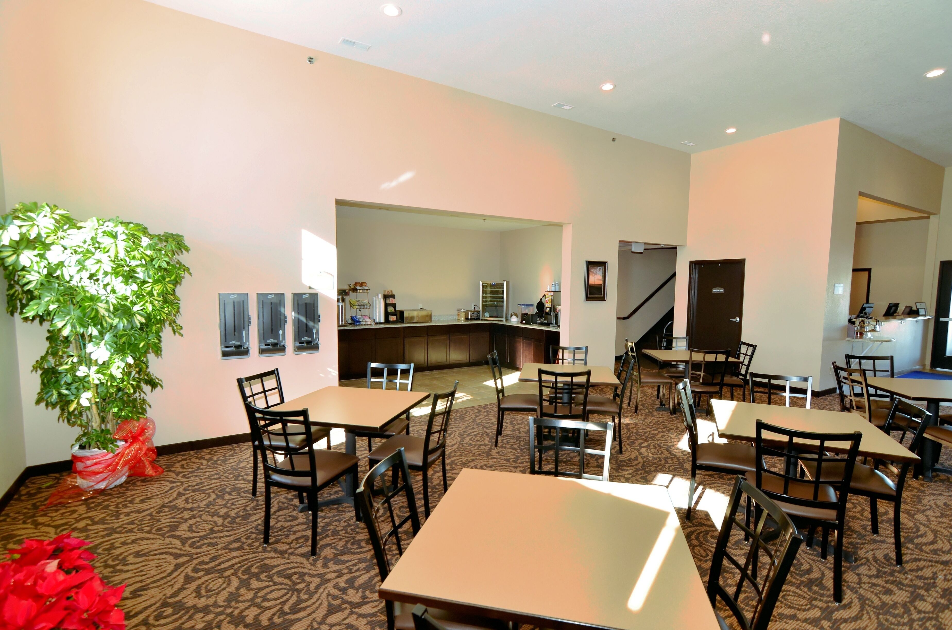 A hotel lobby with multiple tables and chairs, a large potted plant, and a kitchen area in the background.