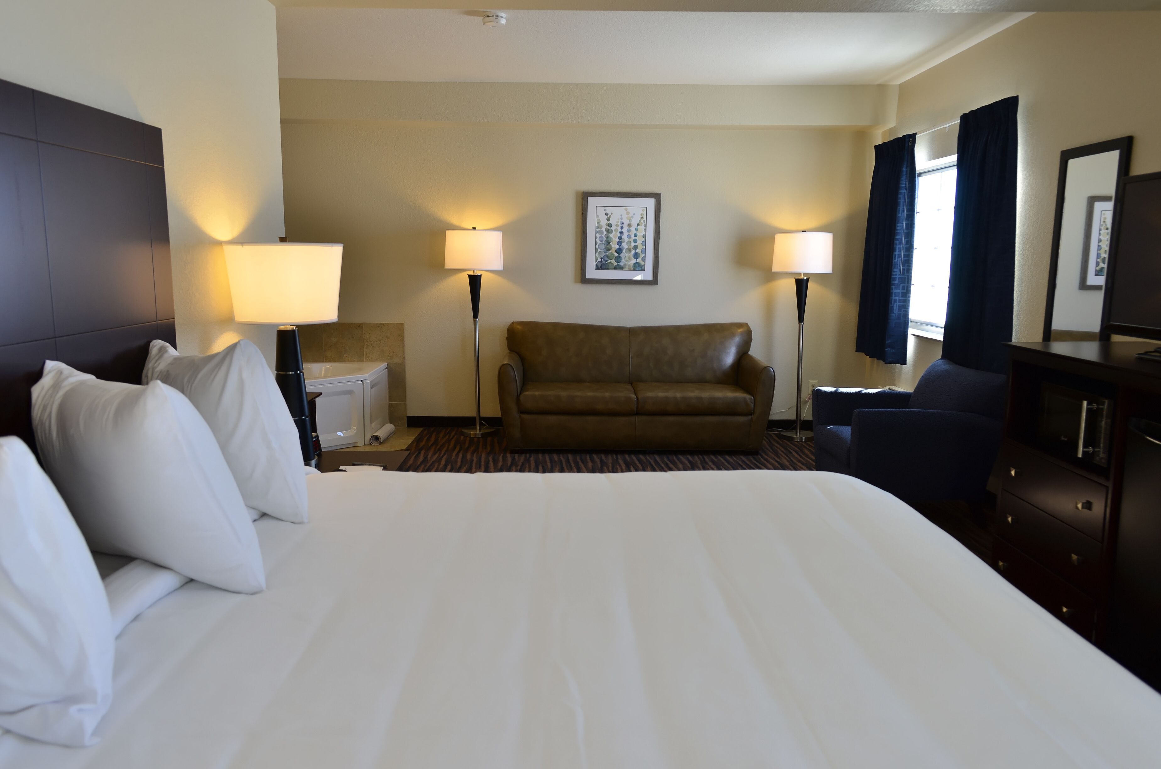 Habitación de hotel con una cama grande, un sofá marrón, dos lámparas de noche, un cuadro enmarcado en la pared y un tocador con espejo.