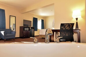 Habitación de hotel con una cama grande, un escritorio con ordenador, una silla, un televisor y un baño con lavabo y espejo.