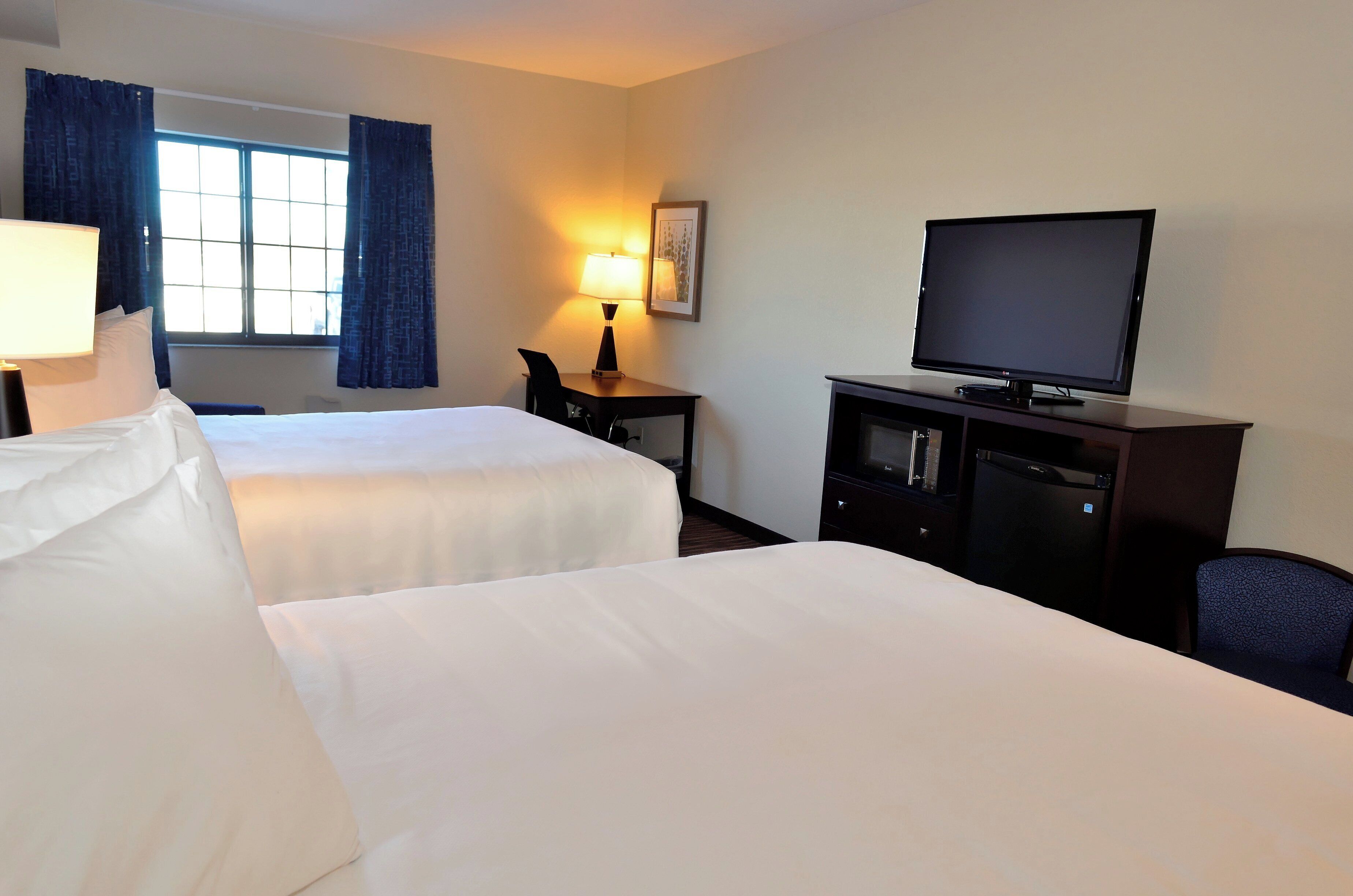A hotel room with two beds, a desk with a lamp, a television, and a window with curtains.