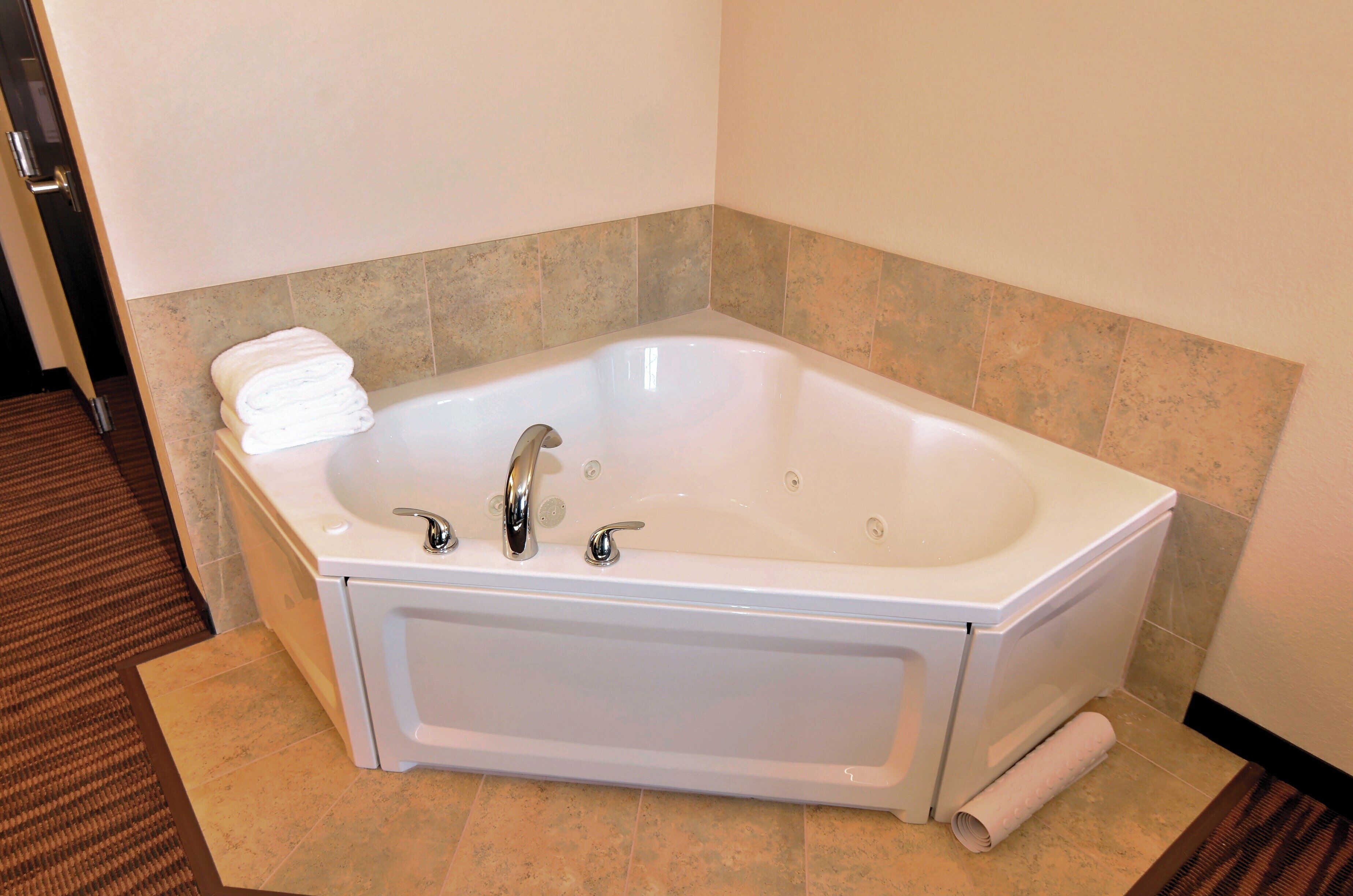 A large, white freestanding bathtub with two faucets and a single control knob, positioned in a corner with beige tiles and a brown carpeted floor.