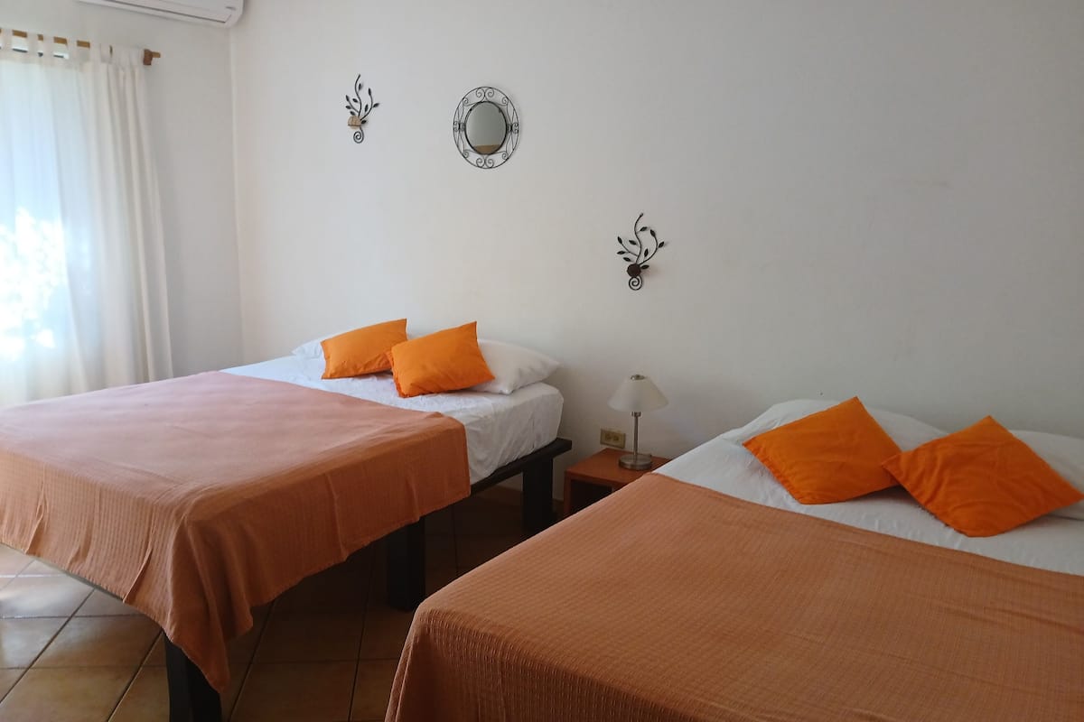 A hotel room with two beds, each with orange bedding and pillows, a bedside table with a lamp, and a wall-mounted mirror.