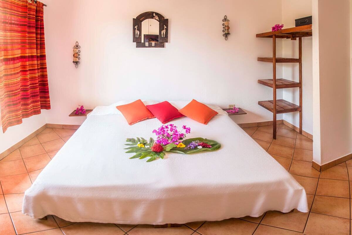 A hotel room with a large bed, orange pillows, a wooden shelf, and a wall-mounted mirror.