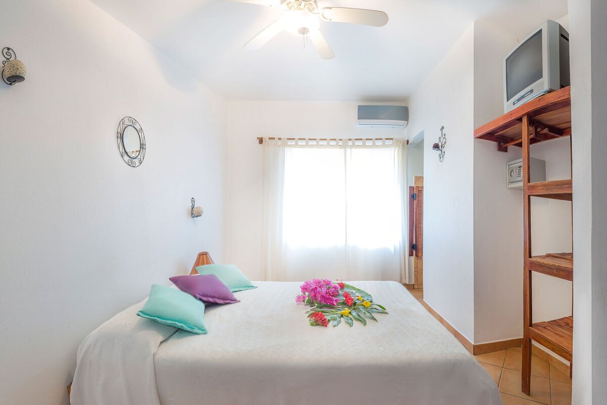 A bedroom with a bed, pillows, a ceiling fan, a TV on a wooden shelf, and a window with curtains.