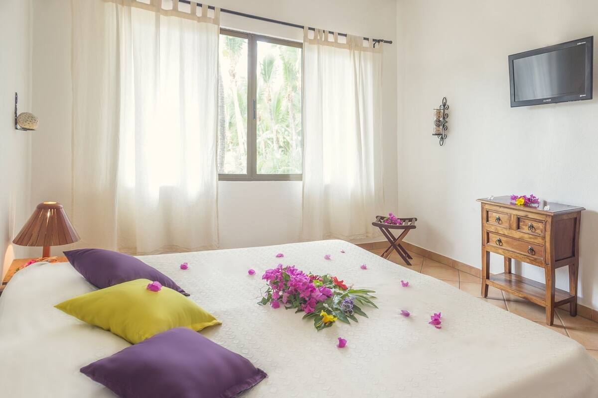 A bedroom with a bed, a nightstand, a lamp, a dresser, a TV, and a window with curtains.