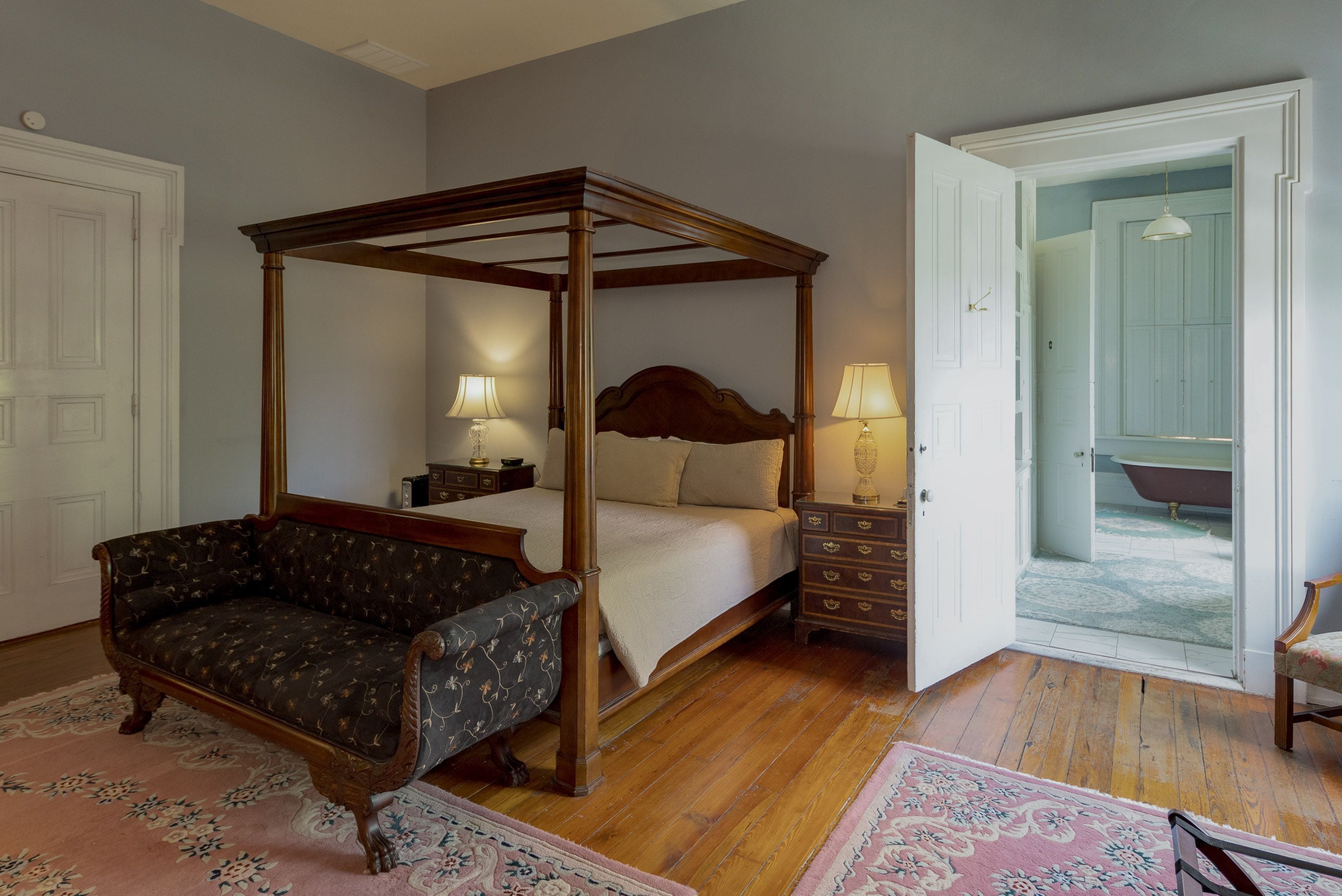 Uma cama com dossel e estrutura de madeira, um sofá, uma mesa de cabeceira com abajur e vista para um banheiro por uma porta aberta.