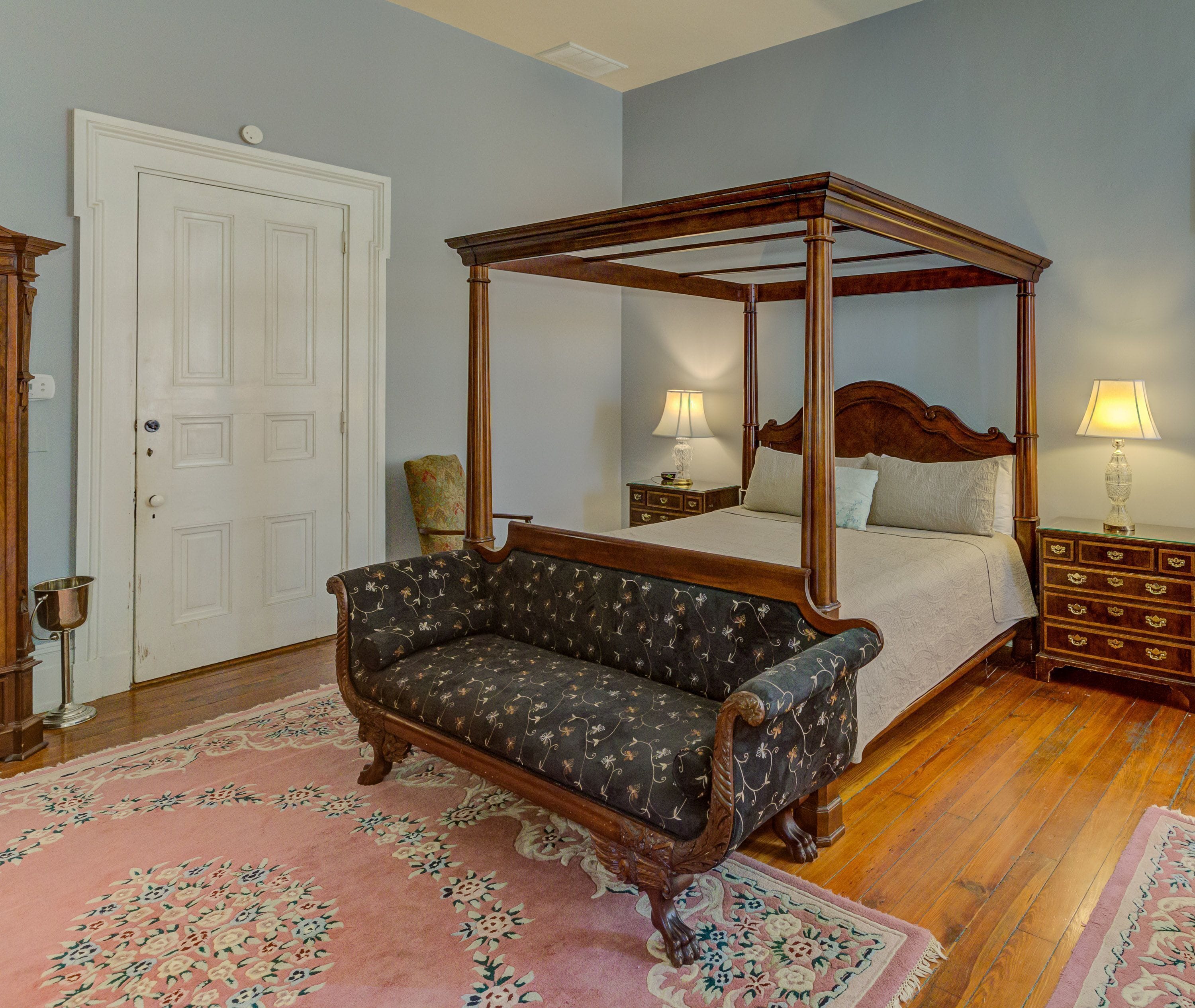 A four-poster canopy bed with a floral-patterned sofa and a pink floral rug in a room with wooden floors and a white door.