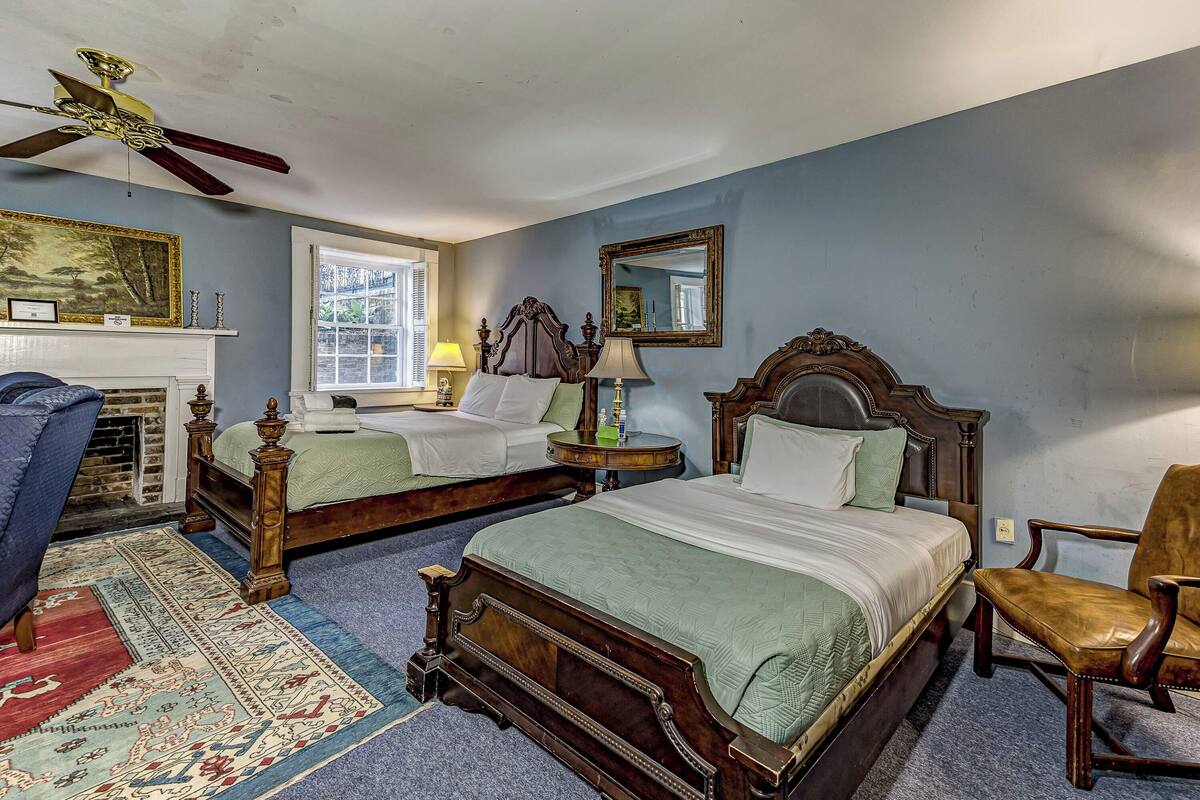 A bedroom with two wooden beds, a ceiling fan, a fireplace, a window, and a chair.