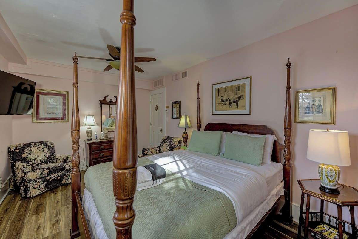 A four-poster bed with a wooden canopy, a nightstand with a lamp, a floral armchair, and framed pictures on the walls.