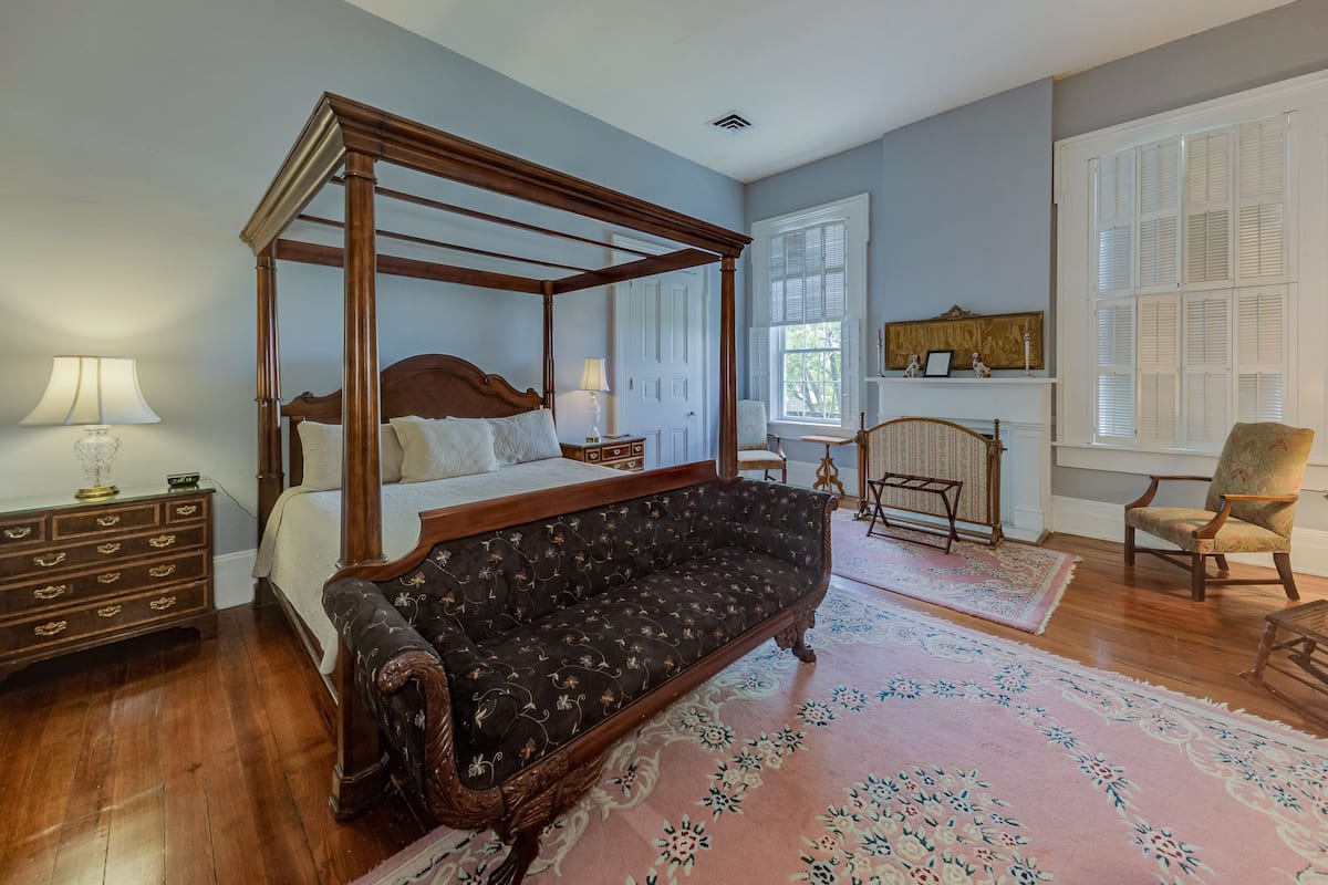 A four-poster bed, a sofa, a chair, and a dresser in a room with wooden floors and a large window.