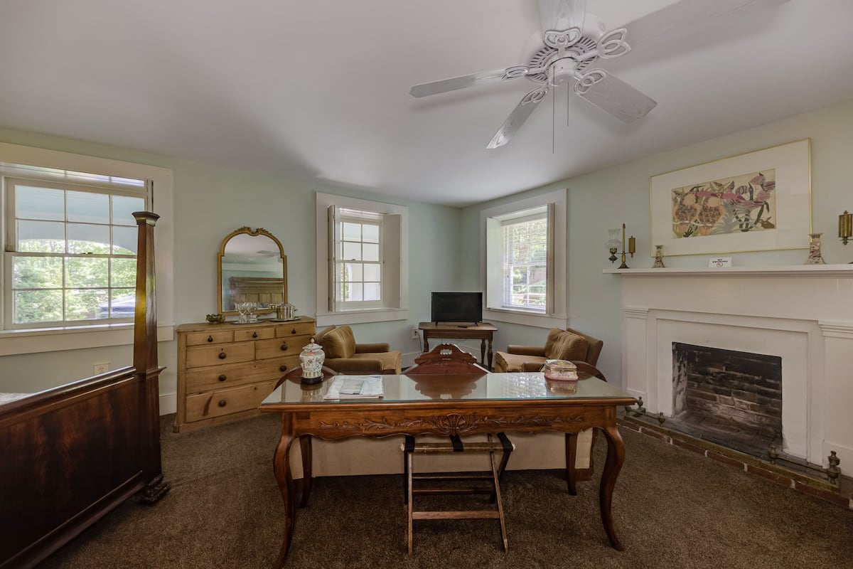 A spacious room with a wooden desk, two chairs, a dresser, a television, and a fireplace.