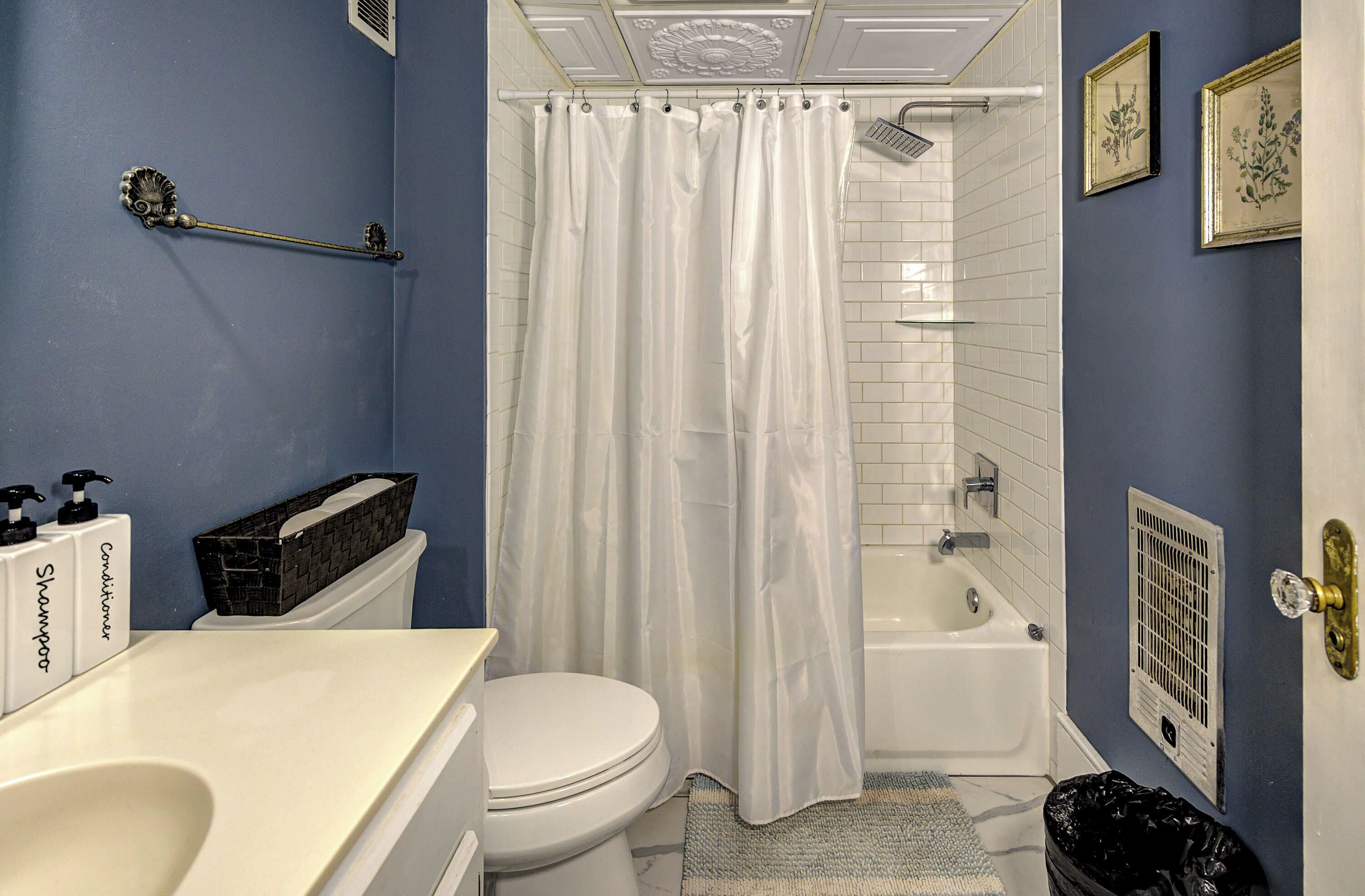 A bathroom with a white toilet, a bathtub, a shower curtain, a sink with a soap dispenser, and a trash bin.
