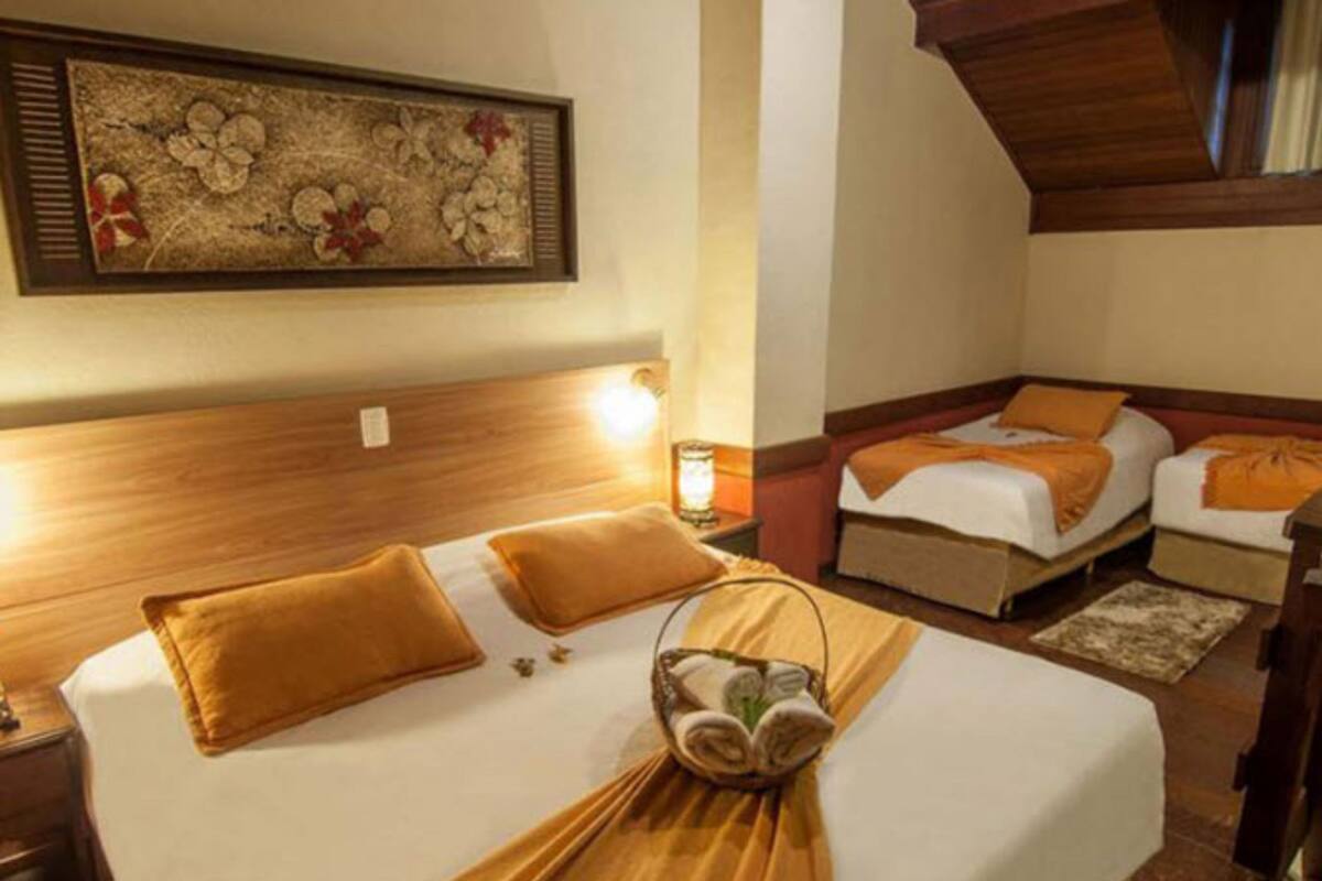 Une chambre d’hôtel avec un grand lit, deux petits lits, une tête de lit en bois, une œuvre d’art encadrée au mur et une table de chevet avec une lampe.