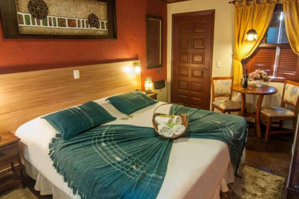 Un lit bien fait, recouvert d’une couverture verte, avec une tête de lit en bois, une œuvre d’art accrochée au mur, une porte en bois, une petite table avec des chaises et une fenêtre ornée de rideaux jaunes.