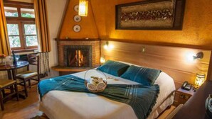 Un dormitorio acogedor con chimenea, una cama con manta azul, una zona de comedor con mesa y sillas, y vistas a la vegetación.