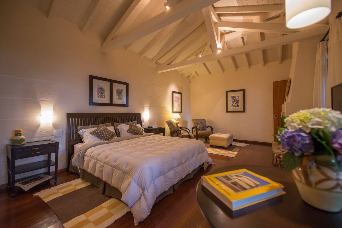 Une chambre avec un grand lit, des tables de chevet, une commode, une chaise, un bureau avec un livre et un vase rempli de fleurs.
