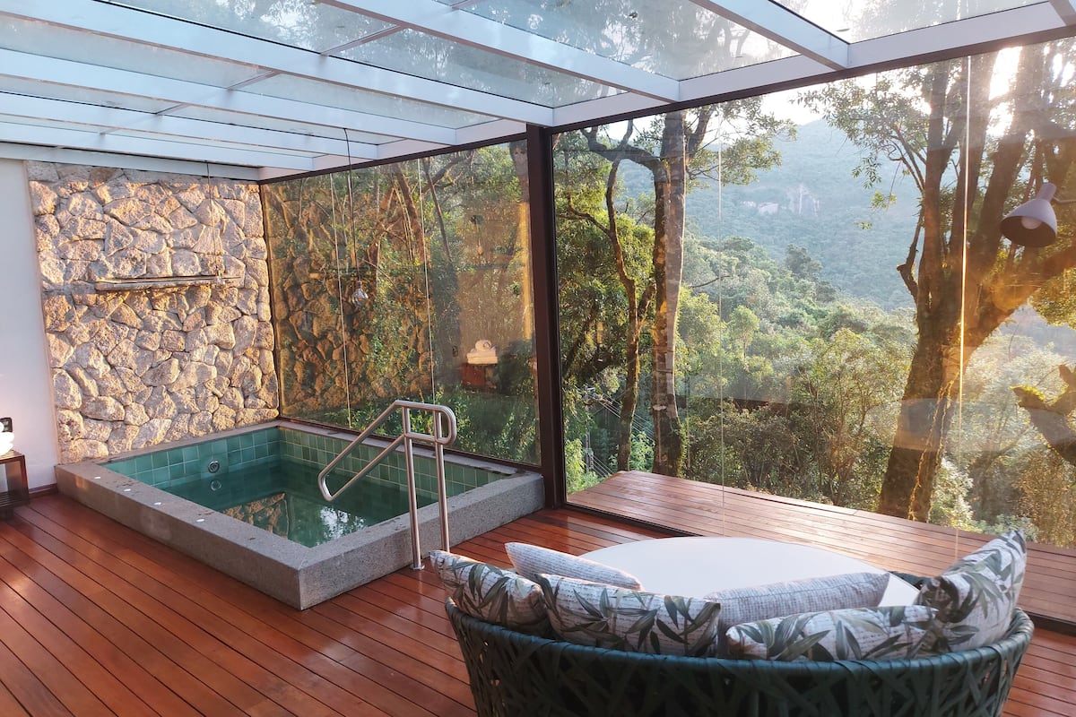 Un espace abrité en verre comprenant un jacuzzi, un mur en pierre, une terrasse en bois et un fauteuil en osier avec des coussins.