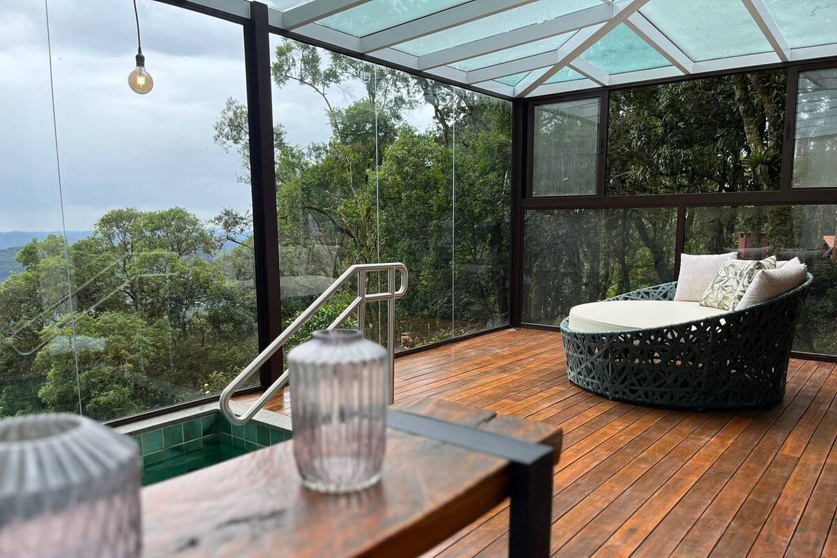 Un espace extérieur moderne, aménagé avec des sièges, une structure en verre, un plancher en bois et offrant une vue sur un paysage boisé.